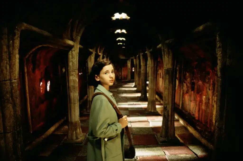 A young girl stands cautiously in a dark, foreboding stone corridor in Guillermo del Toro's Pan's Labyrinth, her wide eyes reflecting both fear and curiosity.