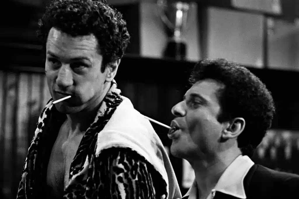 Robert De Niro as boxer Jake LaMotta, wearing a robe, stares intensely with a lollipop in his mouth, next to Joe Pesci, in a gritty black-and-white scene from Raging Bull.