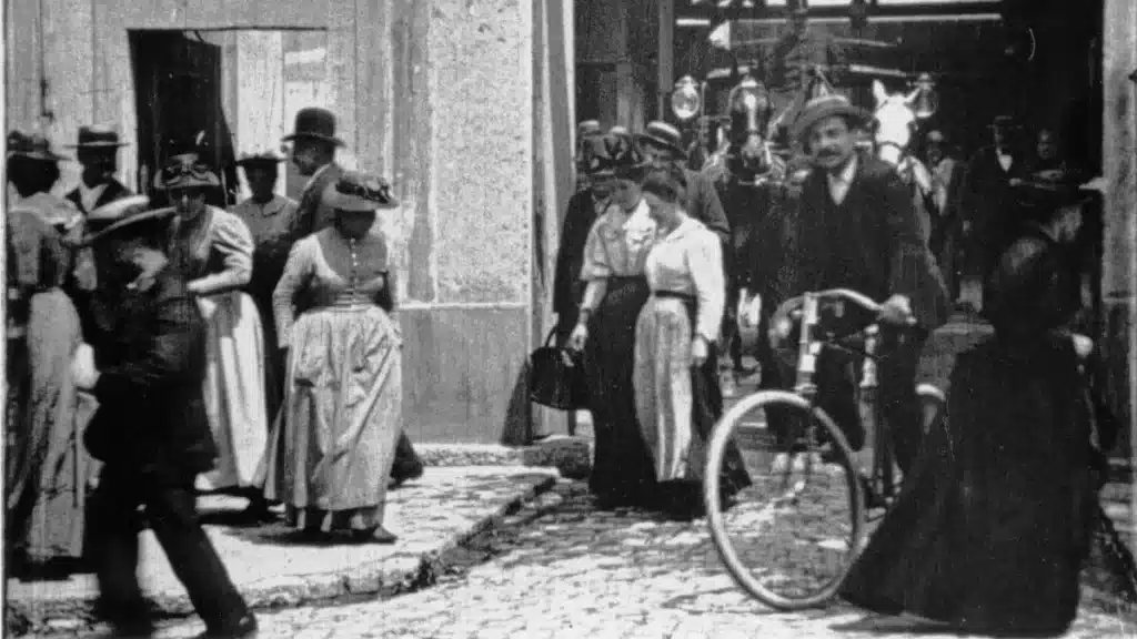 A group of men and women in late 19th-century attire, some wearing hats and dresses, walk out of the Lumière Factory, in one of the earliest recorded films in cinematic history.